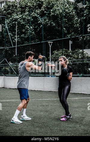 Erwachsene Frau training Muay Thai mit männlichen Trainer im grünen Feld mit Gebäude im Hintergrund Stockfoto