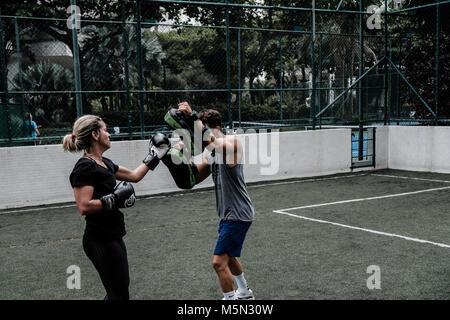 Erwachsene Frau training Muay Thai mit männlichen Trainer im grünen Feld mit Gebäude im Hintergrund Stockfoto