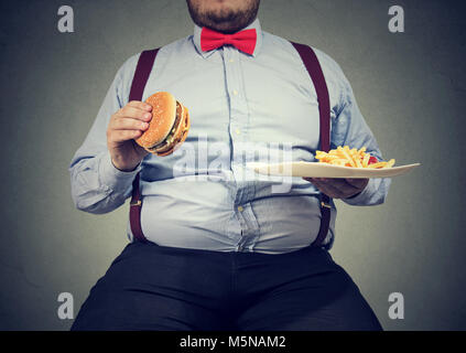 Erntegut Schuß von großer Mann in formelle Kleidung sitzen und konsumieren Platte mit Fast-food auf Grau. Stockfoto