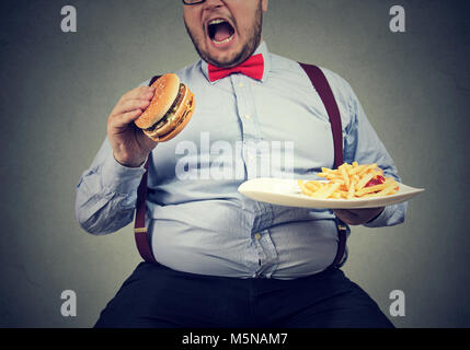 Großer Mann in formelle Kleidung sitzen und konsumieren Platte mit Fast-food auf Grau. Stockfoto