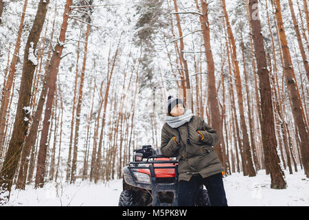 Ein Junge steht in der Nähe ein Quad in der Mitte eines verschneiten Kiefernwald Stockfoto