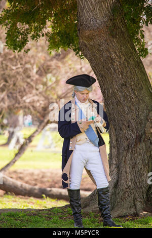 Ein Mann als amerikanische Continental infanterist an einem revolutionären Krieg reenactment im Schatten eines Baumes mit seinen modernen Tag cellphone gekleidet Stockfoto