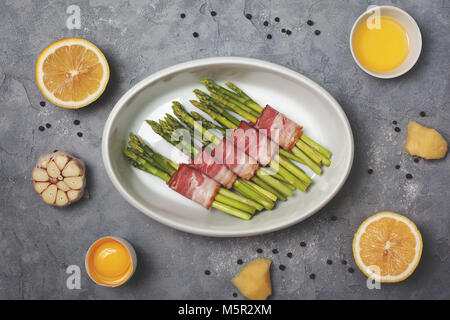 Frischer grüner Spargel mit Schinken in weiße Schüssel zum Backen auf grauem Hintergrund. Ansicht von oben Stockfoto