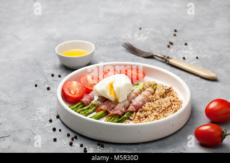 Gesundes Frühstück. Gebackener Spargel mit Schinken, Quinoa, Ei in einer weißen Schüssel auf grauem Stein pochierte Stockfoto