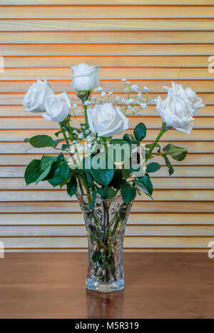 Die Anlage ist ein Blumenstrauß aus den weißen Rosen mit grünen Blättern und kleinen, weißen Blüten, in ein Glas Vase auf einem hölzernen braunen Tisch, vor dem Hintergrund der Ihr Stockfoto