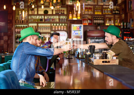 Gruppe von Freunden klirren Biergläser zusammen, während zusammen in der modernen Pub versammelt, um St. Patricks Tag, einige von ihnen tragen Gre zu feiern. Stockfoto