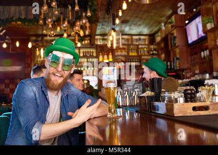 Portrait von fröhlichen rothaarigen Mann, der lustig Party Brille und grüne Melone auf Kamera mit toothy Lächeln beim Sitzen an der Theke, g Stockfoto