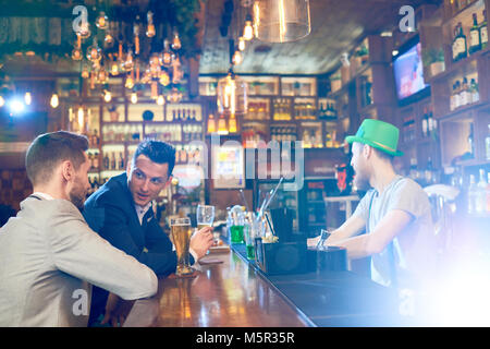 Zwei schöne und weiße Arbeiter tragen klassische Anzüge an der Theke sitzen und plaudern miteinander nach einem harten Arbeitstag, bärtigen Barkeeper w Stockfoto