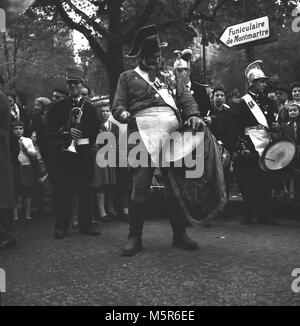 1950, Paris, Frankreich, historische Bild zeigt eine alte französische Soldaten tragen historische Uniformen und Hut, spielt seine Trommel an Montmartre, zusammen mit anderen Mitgliedern einer Militärkapelle. Ein Schild weist in die Richtung der "Seilbahn", das Ski-Lift mit Rädern, die die Menschen an der Spitze des Montmartre Hügels nimmt mit seinen besonderen Blick auf die Stadt. Stockfoto