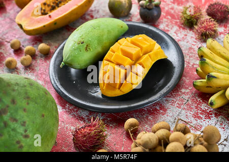 Frische exotische Früchte auf dem schäbigen roten Tisch aus Holz. Es gibt Mangos auf die schwarze Platte, Bananen, Litschis, Rambutan, Papaya pomelo, und mangostanfrüchte. Stockfoto