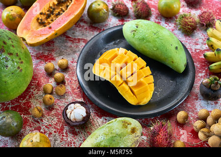 Köstliche exotische Früchte auf dem schäbigen roten Tisch aus Holz. Es gibt Mangos auf die schwarze Platte, Bananen, Litschis, Rambutan, Papaya pomelo, und mangostee Stockfoto