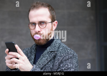 Bärtiger Mann mit Smartphone Blasen einer Blase mit Bubblegum Stockfoto