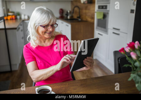 Portrait von gerne ältere Dame am Küchentisch sitzen auf Tablet Stockfoto