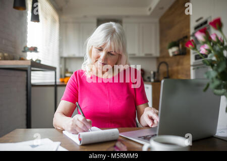 Portrait von älteren Dame am Küchentisch Notizen mit Notebook Stockfoto