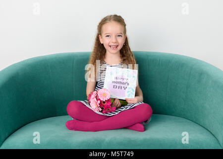 Adorable junges Mädchen sitzt auf einer Couch, Blumenstrauß aus Rosa Gerbera daisies und Muttertag Karte. Happy Mother's Day Konzept. Stockfoto