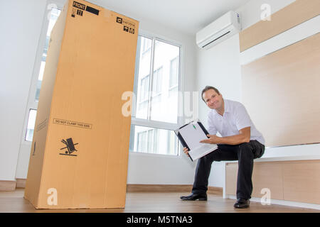 Ein Mann hält Dokumente befindet sich direkt neben einem großen Paket in einem leeren Raum. Der Postbote liefert das Paket in die neue Wohnung. Stockfoto