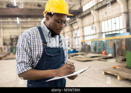 Konzentrierten sich die jungen Techniker tragen insgesamt und Schutzhelm stehen geräumige Produktion moderner Anlagen und Notizen während Stockfoto