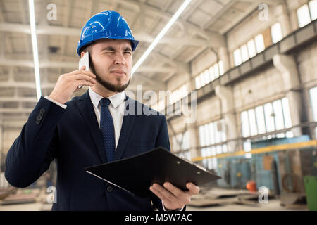 Bärtige junge Unternehmer tragen, hardhat und klassischen Anzug mit seinem Geschäftspartner auf Handy beim Stehen auf geräumige Produktion de Stockfoto