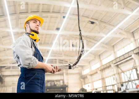 Low Angle View von bärtigen Maschinenbediener tragen insgesamt und Schutzhelm eingehüllt in und stehen Arbeiten im geräumigen Produktion o Stockfoto