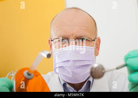 Reife männliche Zahnarzt maskierter Holding zahnmedizinische Instrumente, die Sie in die Kamera schaut Stockfoto