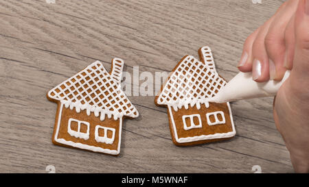 In der Nähe der weiblichen Hand beim Dekorieren süßen Lebkuchen auf Holz Tisch. Die Frau ist die Malerei Haus auf den Keks mit einem sahnehäubchen Tasche mit Zuckerguss. Stockfoto