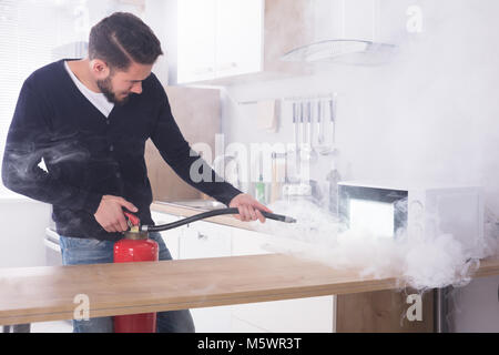 Junger Mann Spritzen Feuerlöscher auf Mikrowelle in der Küche Stockfoto