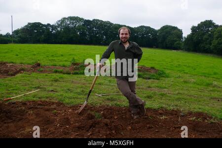Jungen gutaussehenden Mann manuell Graben einer Wiese in Mid Devon zu bauen einen Poly Tunnel. Lydcott Baumschule, Morchard Bishop, Exeter, UK. Agricultura.