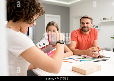 Portrait der aufgeregten Jungen Frau Eröffnung Geschenkbox am Muttertag mit dem Rest der Familie im Hintergrund Stockfoto