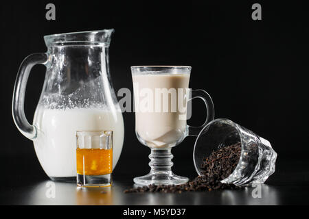 Schwarzer Tee ans Krug Milch aus der Flasche in das Glas auf schwarzem Hintergrund. Stockfoto