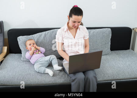 Frau sitzt auf einem Sofa mit Laptop, während Ihr Baby Spielen mit Spielzeug Stockfoto