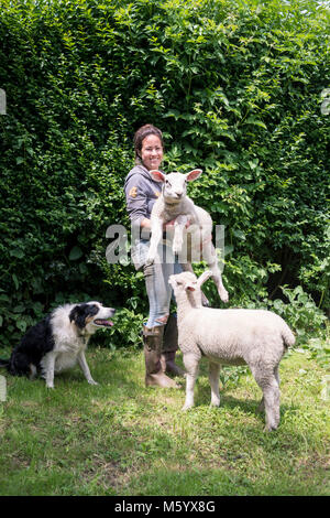 Eine junge weibliche Bauer betreut und versorgt, während mit einigen wachsenden Lämmer in ein Feld auf Ihrem Bauernhof posieren. Stockfoto