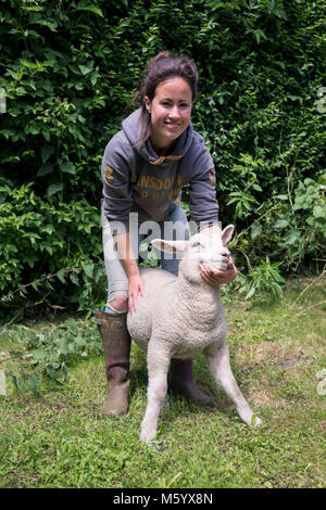 Eine junge weibliche Bauer betreut und versorgt, während mit einigen wachsenden Lämmer in ein Feld auf Ihrem Bauernhof posieren. Stockfoto