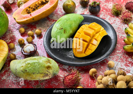 Saftige exotische Früchte auf dem schäbigen roten Tisch aus Holz. Es gibt Mangos auf die schwarze Platte, Bananen, Litschis, Rambutan, Papaya pomelo, und mangostanfrüchte. Stockfoto