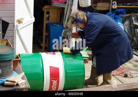 Teenager mit langen blonden Haaren, Laborkittel und Sicherheitsausrüstung Hämmern Löcher in einem gebrauchten Ölfass. DIY-Projekt, Hobo Feuer. Exeter, Devon, Großbritannien. Stockfoto