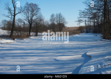 Eis und Schnee bedeckten Fluss, Bäume, Binsen, gefallenen Felder und blauer Himmel mit weißen Wolken. Kalte, verschneite Winterlandschaft. Stockfoto