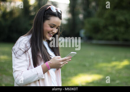 Junge schöne Frau mit Smartphone im Freien in einem Park, etwas zu lesen, interessant und lustig Stockfoto