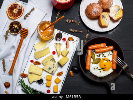 Weißes Holzbrett mit Stücken von verschiedenen Käse und einen runden, gusseisernen Pfanne mit Spiegelei und Würstchen auf einem schwarzen Holztisch, top v Stockfoto