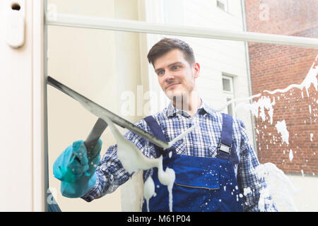 Junge männliche Haushälterin in Uniform Reinigung Fenster mit Rakel Stockfoto