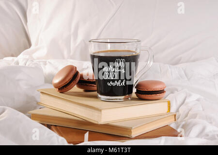 Morgendlichen Tasse Kaffee mit Schokolade Kuchen Makronen, auf einem Stapel Bücher im Bett. Die Inschrift auf dem Becher "Guten Morgen", "Crazy World". Stockfoto