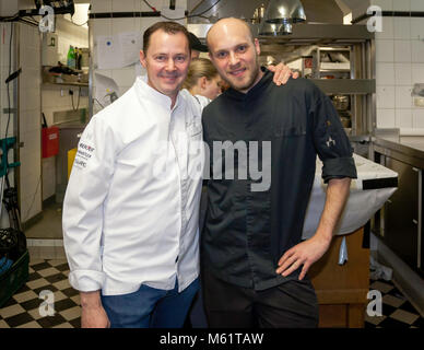 Michelin-Sterne-Köche Simon Taxacher (links) und Simon Stirnal (rechts), der Gastgeberkoch, stehen lächelnd nebeneinander in der Küche des Hotels und Restaurants Kronenschlösschen in Hattenheim, Eltville am Rhein, Hessen, Deutschland. Stirnal legt seinen rechten Arm um Taxachers Schulter und stützt seine linke Hand auf seine Hüfte. Taxacher trägt eine weiße Kochjacke mit Sponsordrucken am Ärmel, Stirnal eine schwarze Kochjacke mit einem Stern am Ärmel. Der Küchenboden hat schwarze und weiße Fliesen. Beim 21. Rheingau Gourmet & Wein Festival kocht Taxacher das "Österreich Dinner" (Österreich-Dinner) am geraden Stockfoto