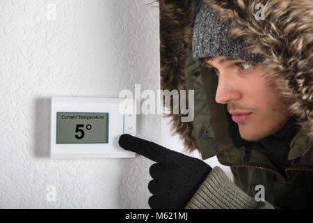 Junger Mann in warme Kleidung, die auf aktuelle Raumtemperatur in Digitaler Thermostat zu Hause Stockfoto