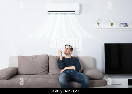 Junge glücklicher Mann auf der Couch, die Klimaanlage mit Fernbedienung zu Hause sitzen Stockfoto