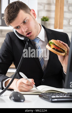 Young Business Mann an Schreibtisch essen Burger und Arbeiten am Arbeitsplatz Stockfoto