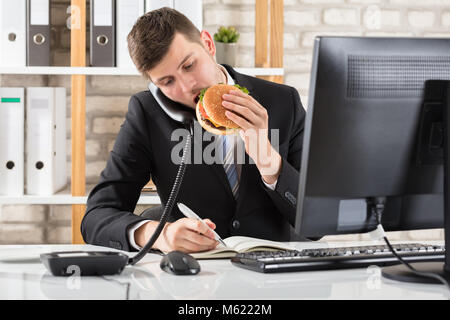 Young Business Mann an Schreibtisch essen Burger und Arbeiten am Arbeitsplatz Stockfoto