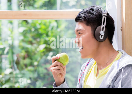 Junger Mann, der ein frisches Grün Apple beim Hören mit Kopfhörern Stockfoto