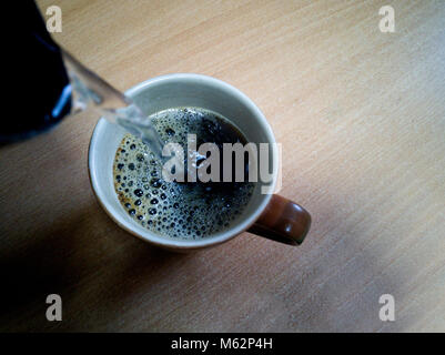 Vorbereitung von Instant Kaffee im Büro. In Tasse mit Kaffee kochendes Wasser aus dem Wasserkocher, Kaffee gießen bildet Schaum. Stockfoto