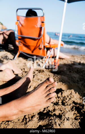 Nahaufnahme der Füße von zwei jungen kaukasischen Männern liegen auf dem Sand des Strandes, und einige unkenntlich Menschen entspannen in Liegestühlen im Hinterg Stockfoto