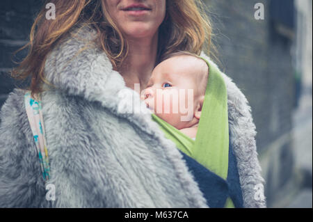 Eine junge Mutter mit ihrem Baby in einem Träger Schlinge steht auf der Straße in der Stadt im Winter Stockfoto