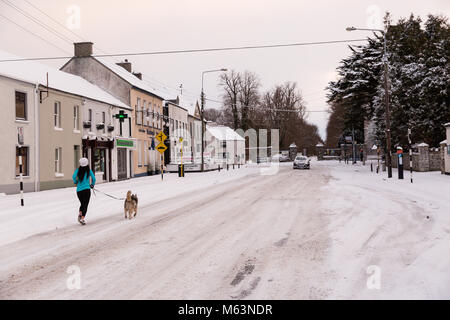 Celbridge, Kildare, Irland. 28 Feb 2018: Irland Wetter. Tier aus dem Osten hits irische Städte. Starker Schneefall in Celbridge. Schnee und Eis sind die Fahrbedingungen erschwert. Frau Joggen mit ihrem Hund auf der Main Street in celbridge. Stockfoto