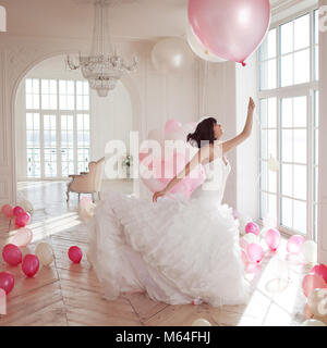 Junge Frau in Wedding Dress in luxuriösen Innenraum fliegt auf rosa und weißen Luftballons. Charmante junge Braut Brünette mit kurzen Haarschnitt im stilvollen Quincean Stockfoto